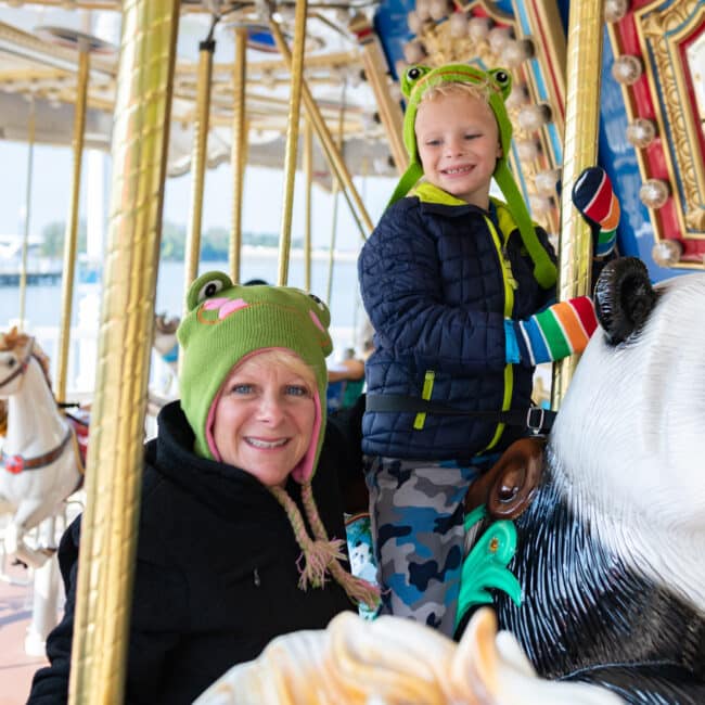 Smiling adult and child riding the Spirit Park Carousel outdoors on a sunny day; child wears green frog hat and stands behind a panda figure, adult wears matching frog hat in foreground, with water and bridge visible in background.